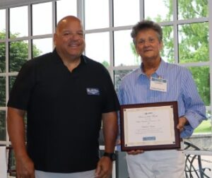 A man and woman ,standing side by side in front or a large multi-paned window, are smiling at the camera while she holds a mounted Business of the Month certificate for Employ-Ease.